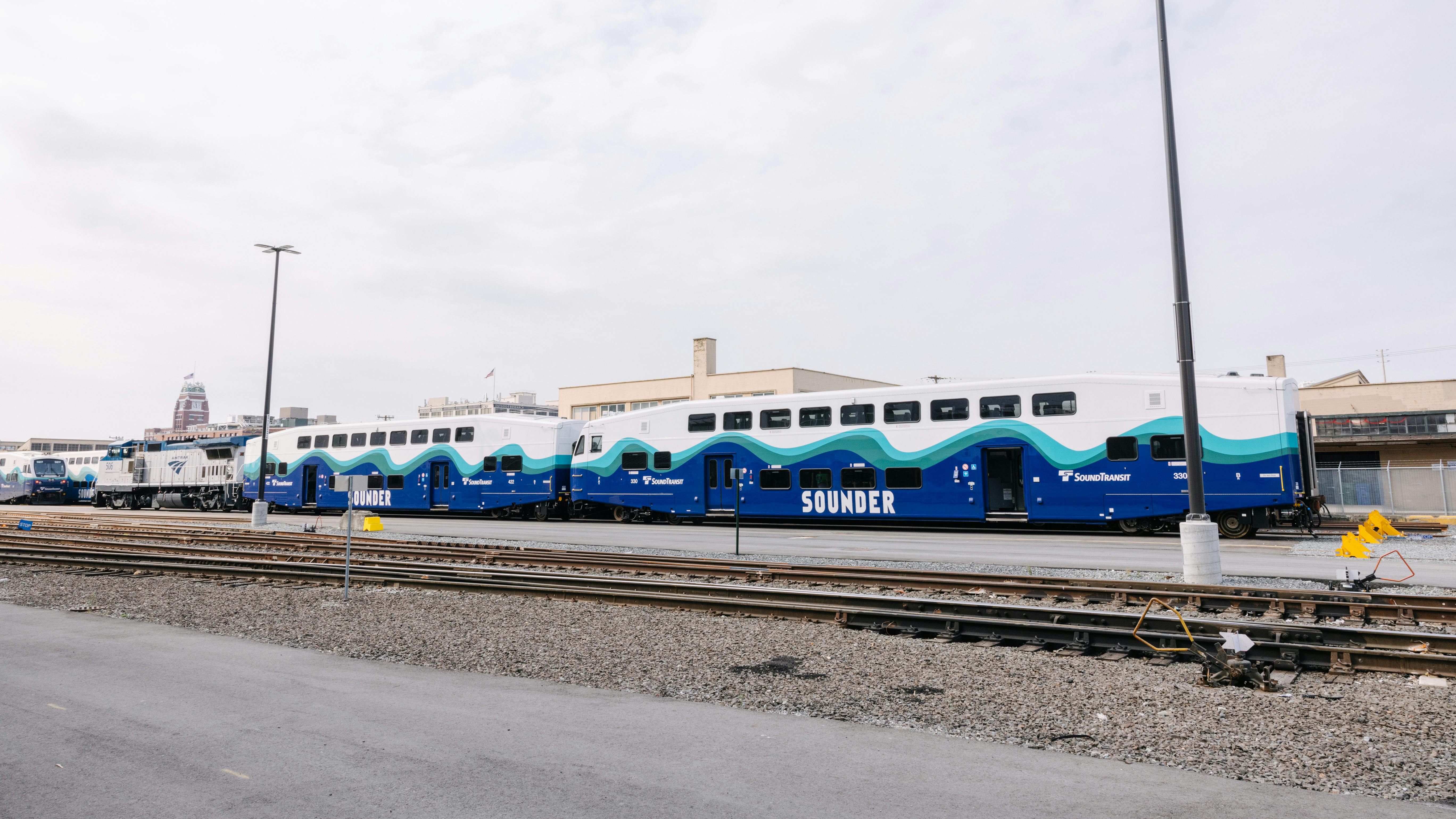 Two new Sound Transit Sounder bilevel commuter rail cars in blue, teal and white wave-pattern livery sit on a rail yard track, with an Amtrak locomotive and additional Sounder equipment visible to the left.