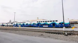 Two new Sound Transit Sounder bilevel commuter rail cars in blue, teal and white wave-pattern livery sit on a rail yard track, with an Amtrak locomotive and additional Sounder equipment visible to the left.