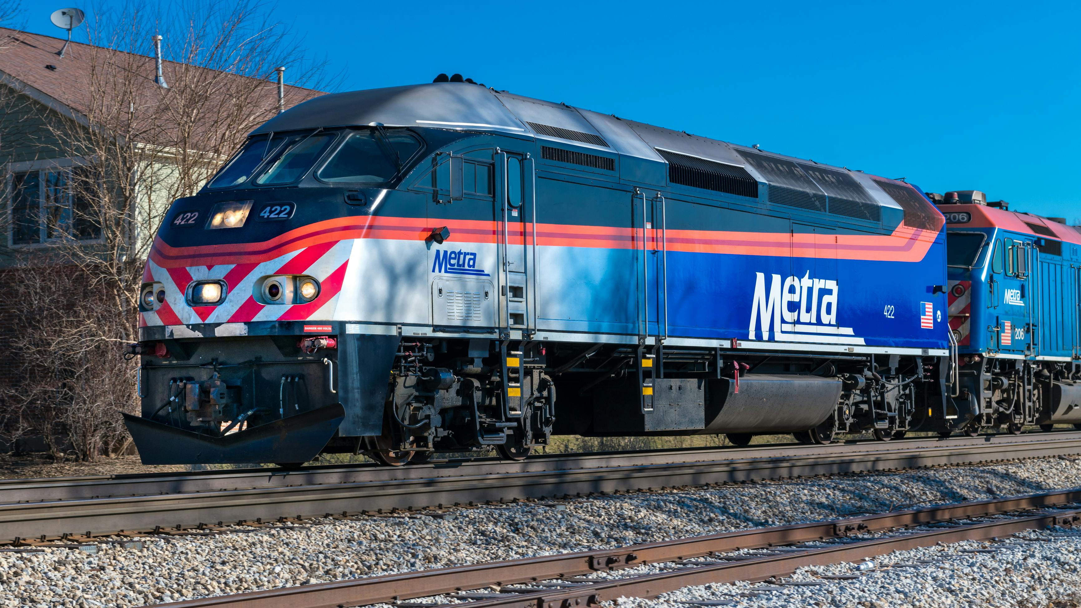 A Metra commuter rail locomotive numbered 422 in the agency's blue, silver and orange livery sits on track in front of a second Metra locomotive, with bare trees and a building behind under a clear blue sky.