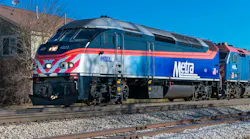 A Metra commuter rail locomotive numbered 422 in the agency's blue, silver and orange livery sits on track in front of a second Metra locomotive, with bare trees and a building behind under a clear blue sky.