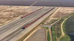An aerial view of the California High-Speed Rail Authority's Central Valley construction site, showing rows of red ballast hopper cars staged alongside parallel sections of prepared trackbed and rail awaiting installation, with surrounding farmland and an irrigation channel to the right. An aerial view of the California High-Speed Rail Authority's Central Valley construction site, showing rows of red ballast hopper cars staged alongside parallel sections of prepared trackbed and rail awaiting installation, with surrounding farmland and an irrigation channel to the right.