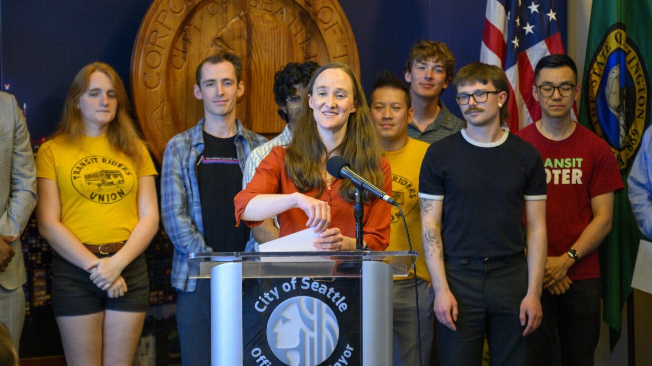 Seattle Mayor Katie Wilson speaks at a podium bearing the City of Seattle Office of the Mayor seal at an event announcing the advancement of the Seattle Transit Measure, flanked by supporters wearing Transit Riders Union and Transit Voter T-shirts, with U.S. and Washington state flags and the carved city seal in the background.