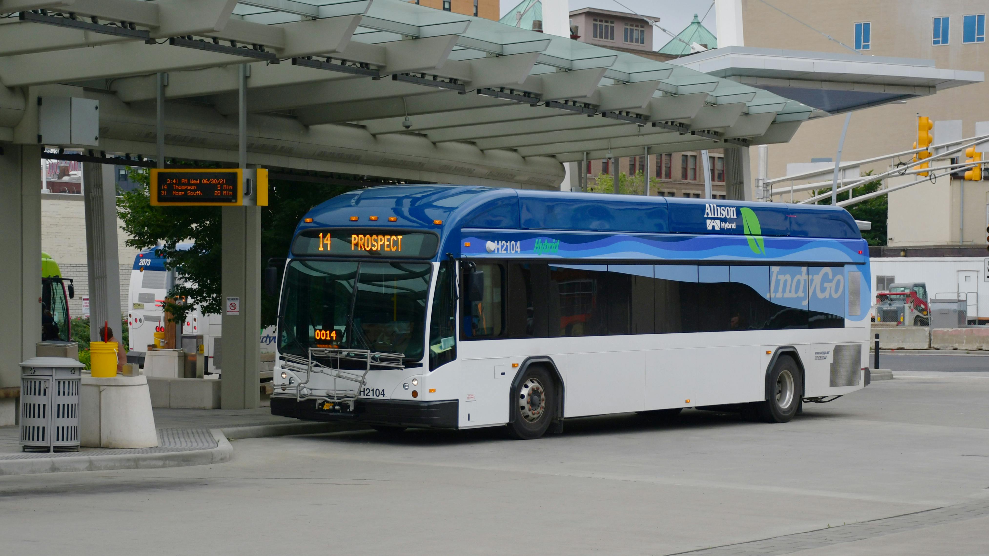 A blue and white IndyGo Allison hybrid bus operating Route 14 to Prospect pulls into a covered downtown transit center platform, with a real-time arrivals sign showing upcoming Routes 14 and 31 and another IndyGo bus visible at an adjacent berth.