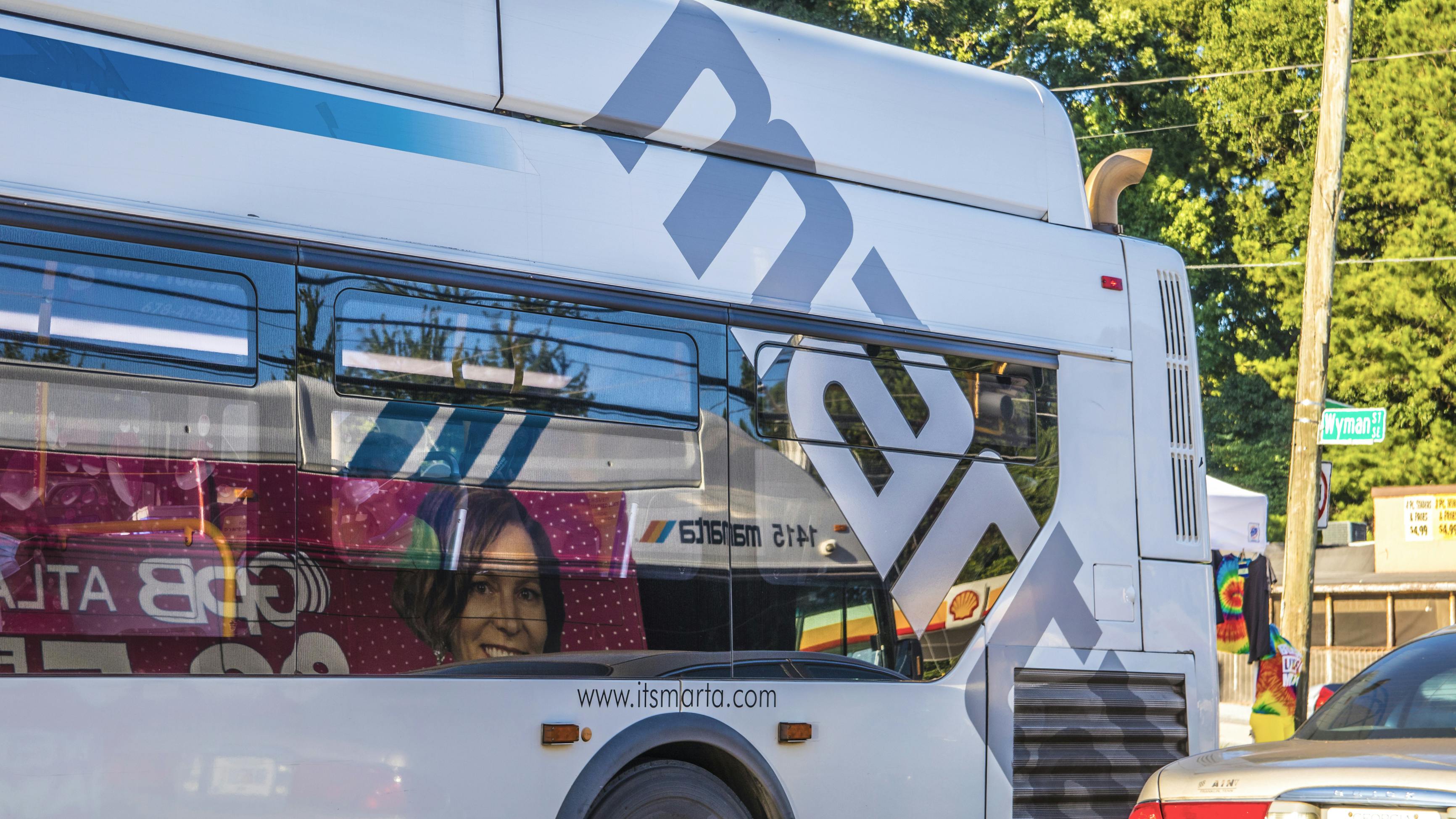A white Metropolitan Atlanta Rapid Transit Authority bus with the agency's gray 'MARTA' logo wrap travels along an Atlanta street, with a passenger visible inside near a interior advertisement and a Wyman Street sign at right.