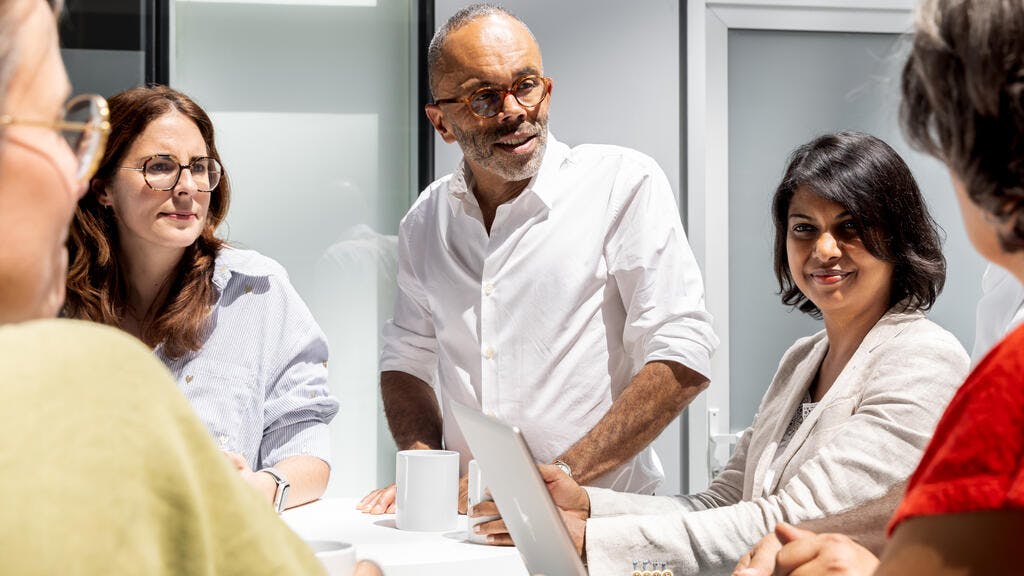 Five Alstom colleagues gather around a conference table in a bright office, with one man standing at the head holding a tablet as he speaks to the seated group over coffee cups.