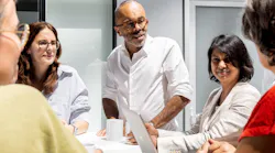 Five Alstom colleagues gather around a conference table in a bright office, with one man standing at the head holding a tablet as he speaks to the seated group over coffee cups.