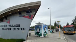 A free-standing Throne self-cleaning public restroom unit sits on the sidewalk at King County Metro's Aurora Village Transit Center, with the center's curved entrance signage at left and a red and yellow RapidRide E Line bus to downtown Seattle at a stop on the right.