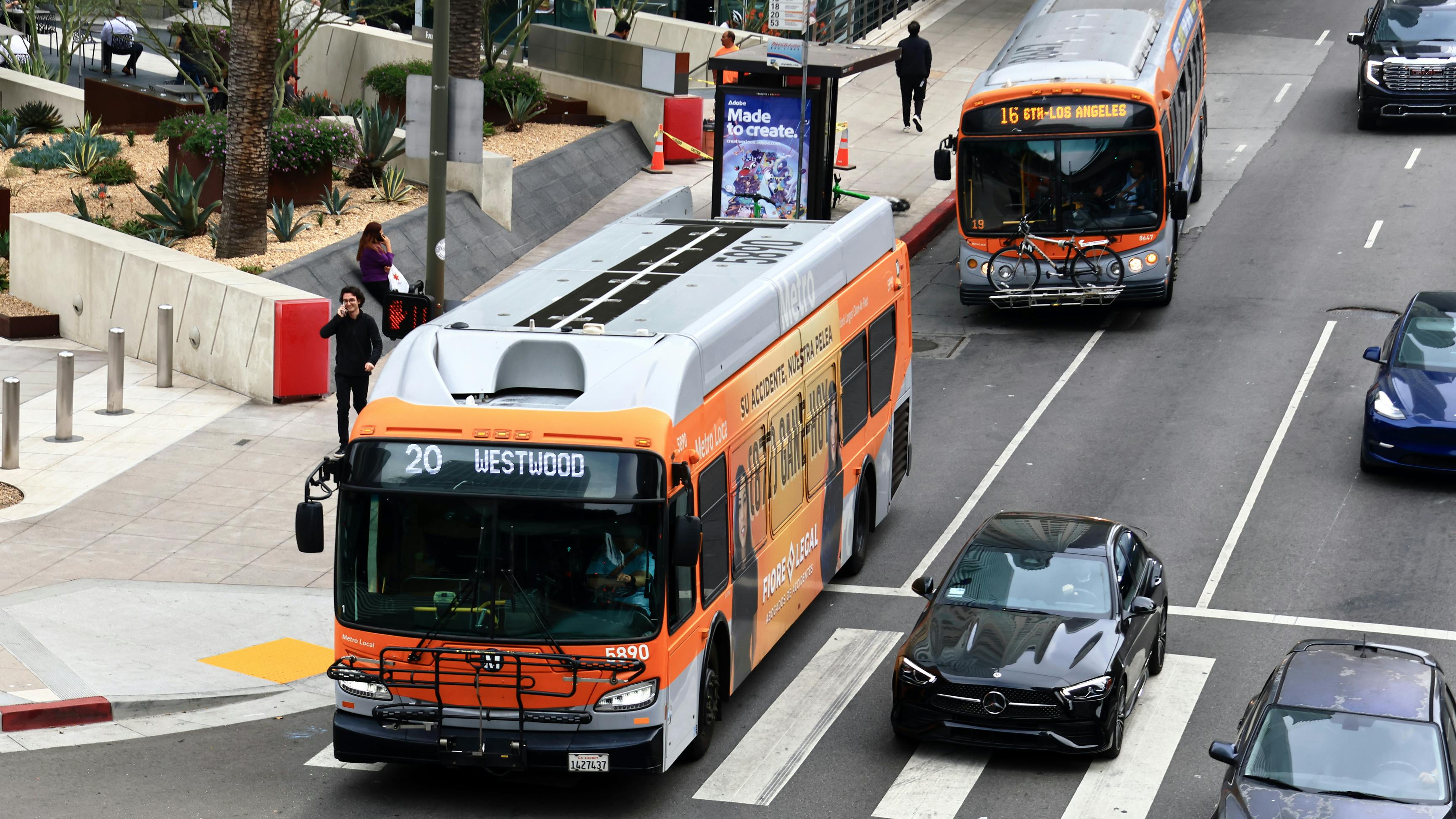 Two orange and silver LA Metro Local buses operate in downtown Los Angeles, with a Route 20 bus to Westwood stopped at a crosswalk in the foreground and a Route 16 bus to 6th-Los Angeles pulling up to a stop in the background, alongside cars on a multi-lane street.