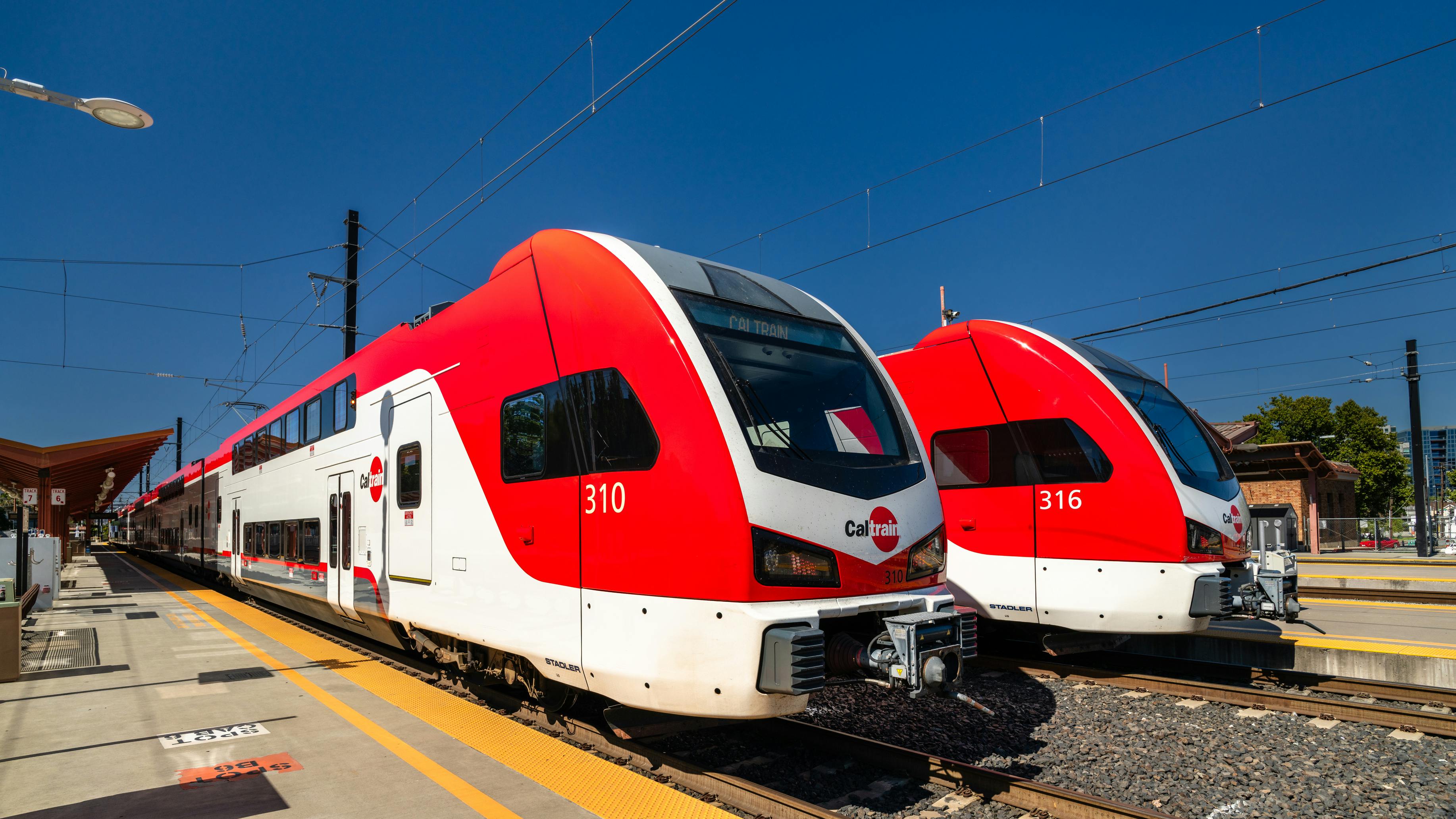 Two Caltrain trains sit next to each other at a station against a clear blue sky.