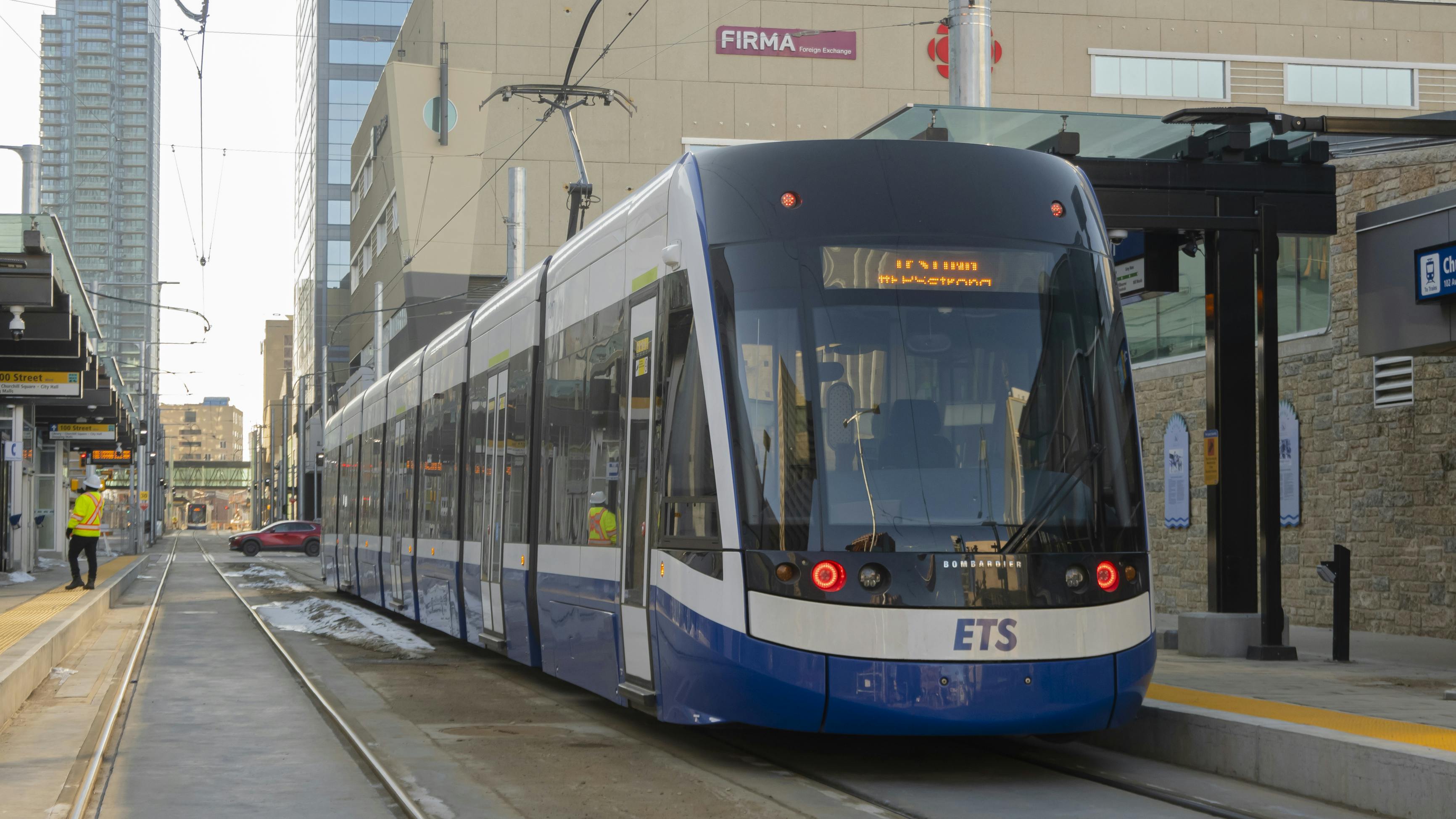A blue and white Edmonton Transit Service Bombardier Flexity light rail vehicle pulls into a downtown Valley Line station, with workers in high-visibility vests on the platform and Edmonton's downtown skyline visible behind.