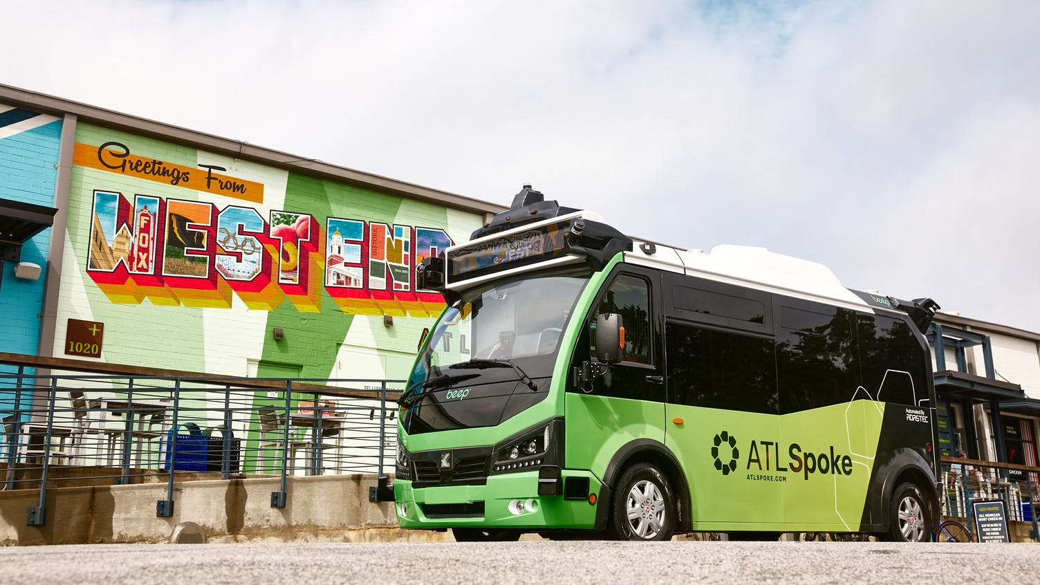 A green ATLSpoke autonomous shuttle from Beep sits parked in front of a colorful 'Greetings From West End' postcard-style mural in Atlanta's Westside neighborhood, with rooftop sensors and lidar equipment visible on the vehicle.