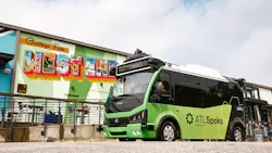 A green ATLSpoke autonomous shuttle from Beep sits parked in front of a colorful 'Greetings From West End' postcard-style mural in Atlanta's Westside neighborhood, with rooftop sensors and lidar equipment visible on the vehicle. A green ATLSpoke autonomous shuttle from Beep sits parked in front of a colorful 'Greetings From West End' postcard-style mural in Atlanta's Westside neighborhood, with rooftop sensors and lidar equipment visible on the vehicle.