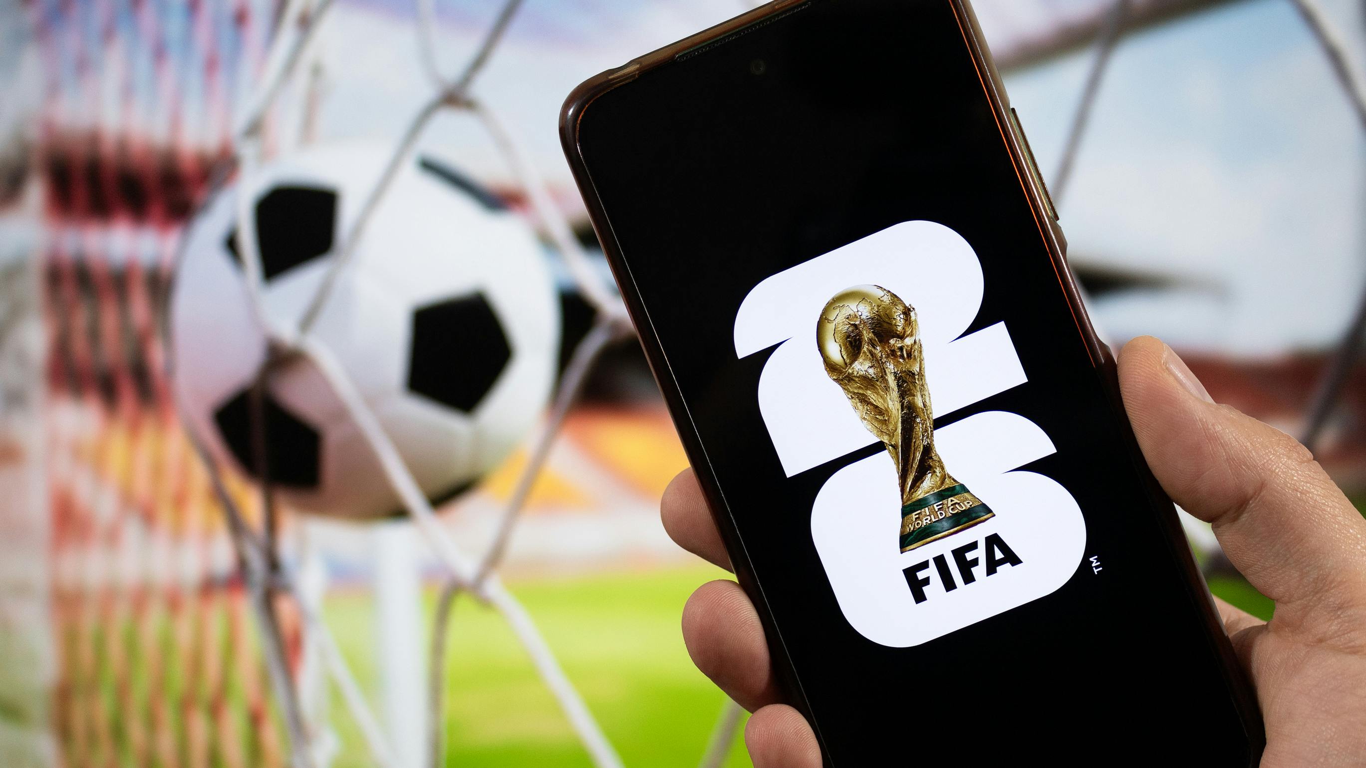 A male hand holding a smartphone with the FIFA World Cup 2026 logo over a black background and a goal blurred background.