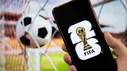 A male hand holding a smartphone with the FIFA World Cup 2026 logo over a black background and a goal blurred background. A male hand holding a smartphone with the FIFA World Cup 2026 logo over a black background and a goal blurred background.