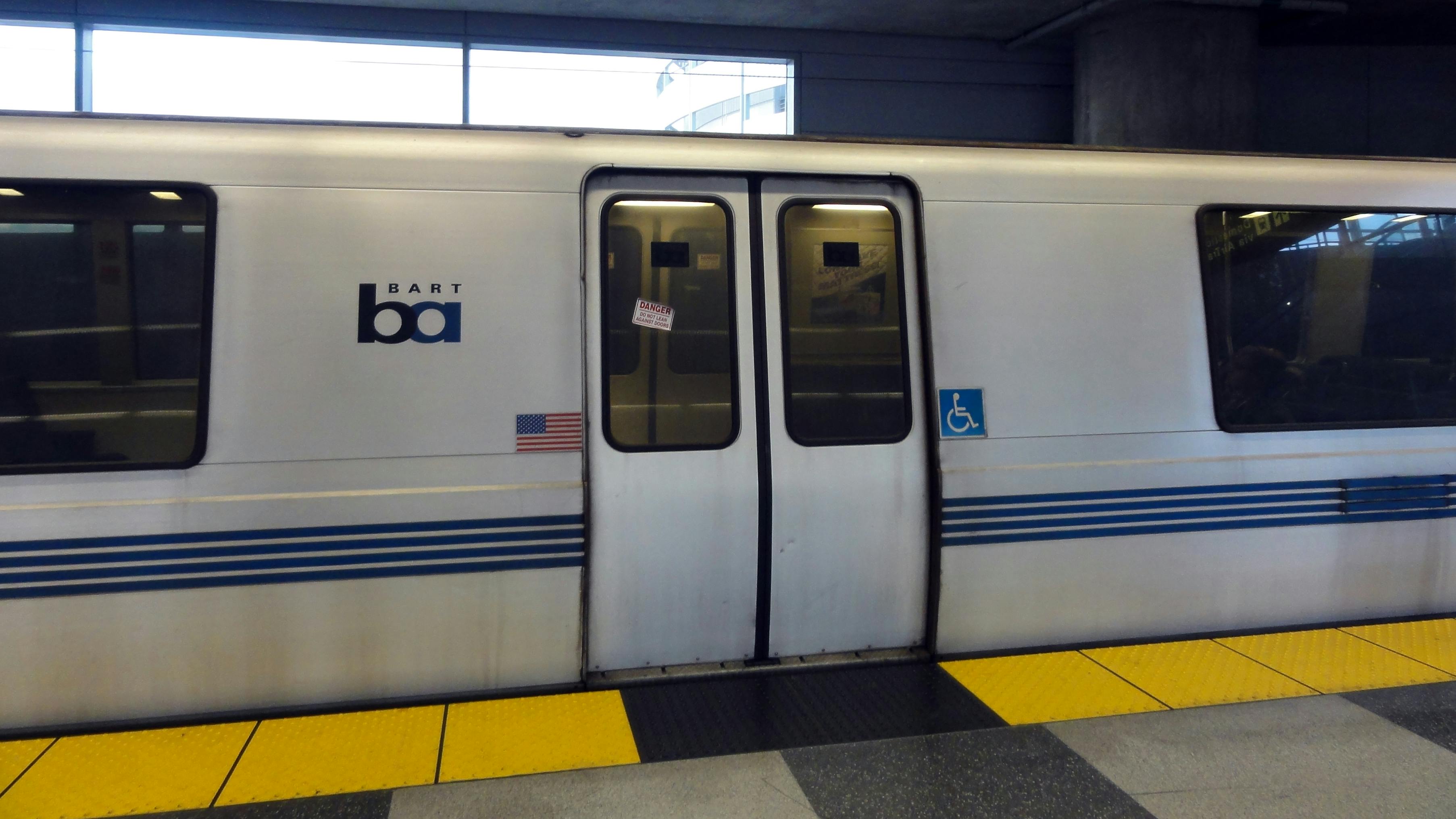 A white Bay Area Rapid Transit train with blue stripes and the BART logo sits at an underground station platform, with closed double doors marked with 'Do Not Lean Against Doors' warnings and accessibility symbols, and yellow tactile warning strips along the platform edge.