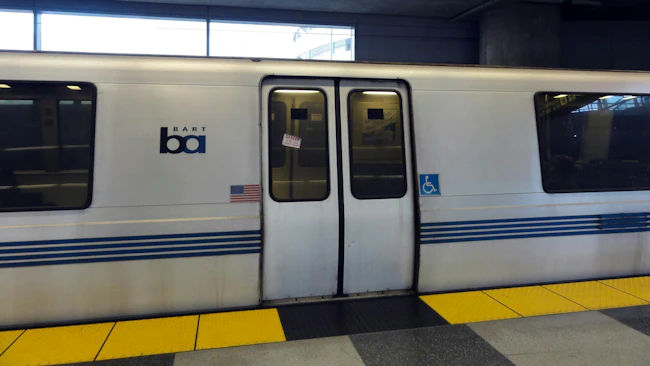 A white Bay Area Rapid Transit train with blue stripes and the BART logo sits at an underground station platform, with closed double doors marked with 'Do Not Lean Against Doors' warnings and accessibility symbols, and yellow tactile warning strips along the platform edge.