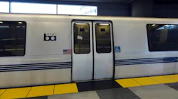 A white Bay Area Rapid Transit train with blue stripes and the BART logo sits at an underground station platform, with closed double doors marked with 'Do Not Lean Against Doors' warnings and accessibility symbols, and yellow tactile warning strips along the platform edge.