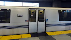 A white Bay Area Rapid Transit train with blue stripes and the BART logo sits at an underground station platform, with closed double doors marked with 'Do Not Lean Against Doors' warnings and accessibility symbols, and yellow tactile warning strips along the platform edge. A white Bay Area Rapid Transit train with blue stripes and the BART logo sits at an underground station platform, with closed double doors marked with 'Do Not Lean Against Doors' warnings and accessibility symbols, and yellow tactile warning strips along the platform edge.