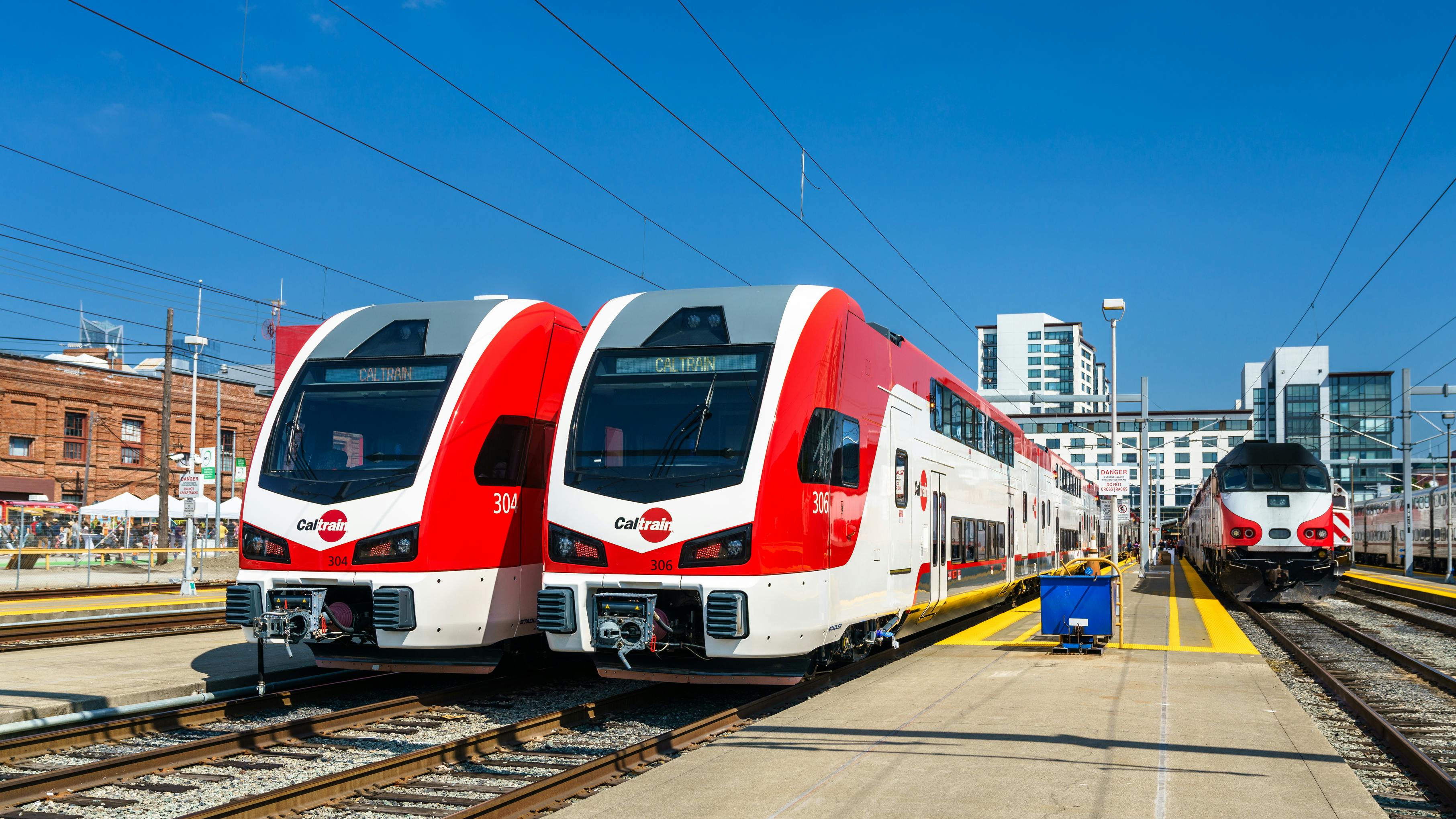 Two new red and white Caltrain Stadler KISS bilevel electric multiple-unit trainsets sit at adjacent platform tracks in San Francisco, with an older Caltrain diesel cab car visible on a third track to the right and downtown buildings in the background under a clear sky.