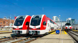 Two new red and white Caltrain Stadler KISS bilevel electric multiple-unit trainsets sit at adjacent platform tracks in San Francisco, with an older Caltrain diesel cab car visible on a third track to the right and downtown buildings in the background under a clear sky.