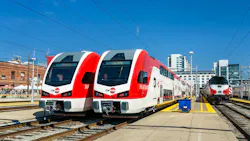 Two new red and white Caltrain Stadler KISS bilevel electric multiple-unit trainsets sit at adjacent platform tracks in San Francisco, with an older Caltrain diesel cab car visible on a third track to the right and downtown buildings in the background under a clear sky. Two new red and white Caltrain Stadler KISS bilevel electric multiple-unit trainsets sit at adjacent platform tracks in San Francisco, with an older Caltrain diesel cab car visible on a third track to the right and downtown buildings in the background under a clear sky.