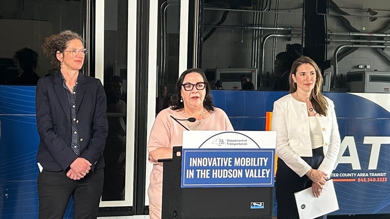 Officials stand on a podium stage in front of a blue Westchester County Area Transit (Bee-Line) bus, with one speaking at a podium bearing a New York State Department of Transportation sign reading 'Innovative Mobility in the Hudson Valley.