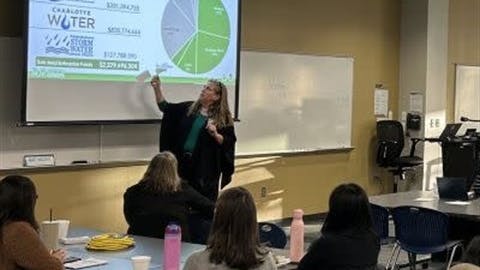 A presenter gestures toward a projected slide showing the Charlotte Area Transit System and other city enterprise funds for fiscal year 2026, with a pie chart breaking down allocations across CATS, Charlotte Water and Storm Water, as attendees take notes in a conference room.