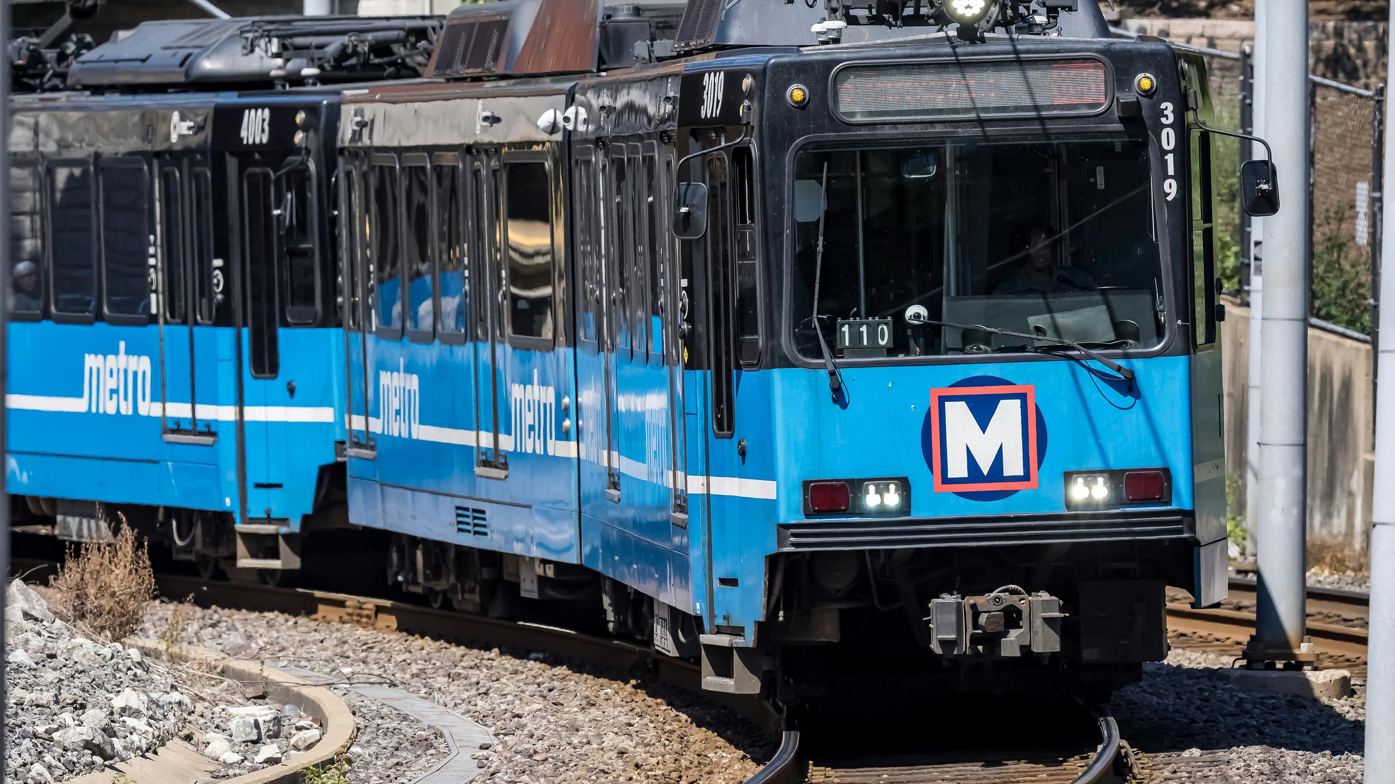 Two coupled blue and white St. Louis Metro MetroLink light rail vehicles, numbered 3019 and 4003, round a curve along the system's at-grade tracks, with overhead catenary wires and a stone retaining wall behind.