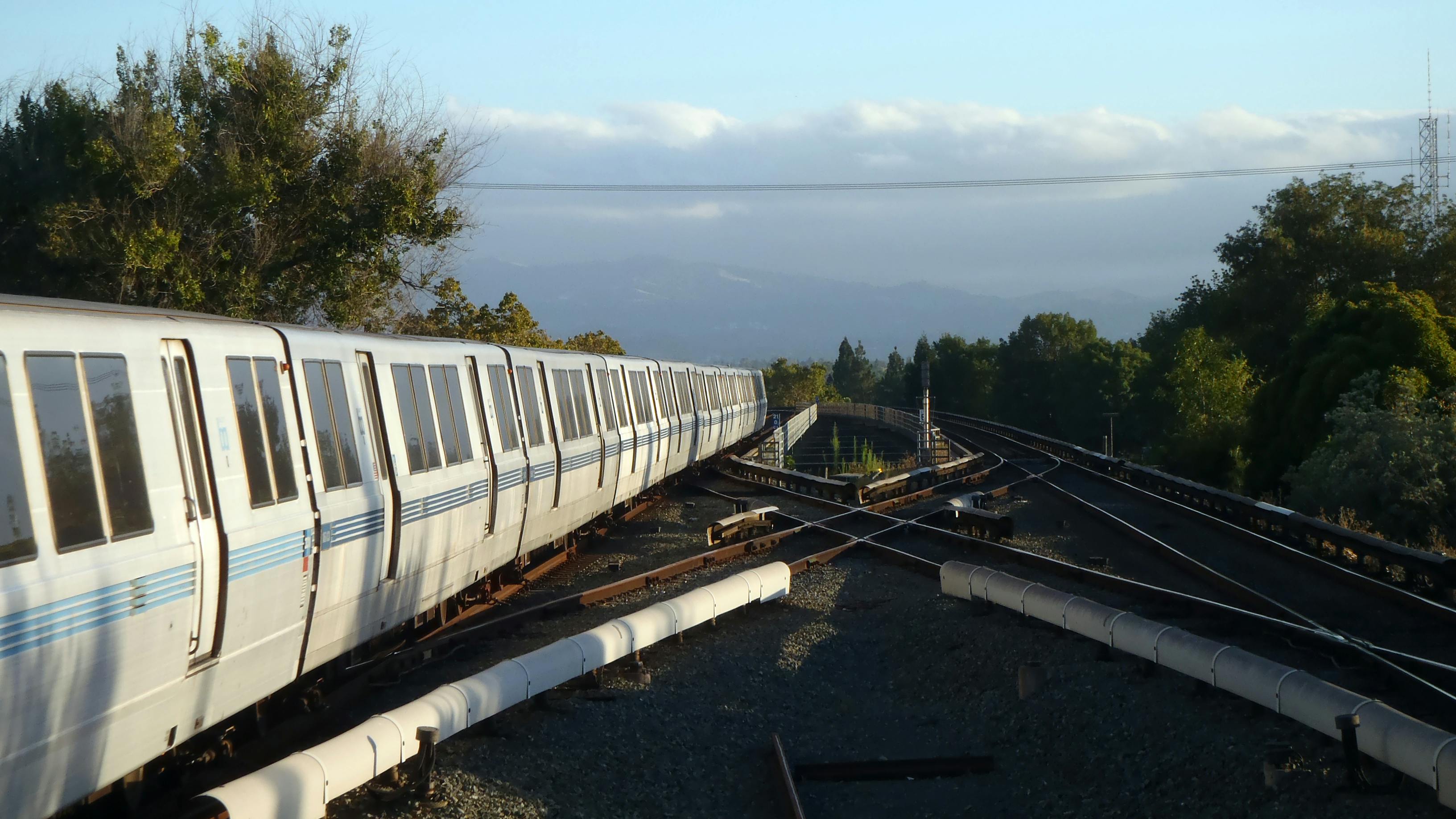 A white and blue Bay Area Rapid Transit train rounds a curve along an elevated section of track, with rolling hills, trees and a partly cloudy sky in the background.