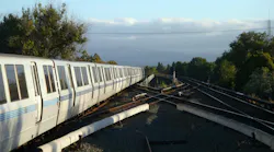 A white and blue Bay Area Rapid Transit train rounds a curve along an elevated section of track, with rolling hills, trees and a partly cloudy sky in the background.