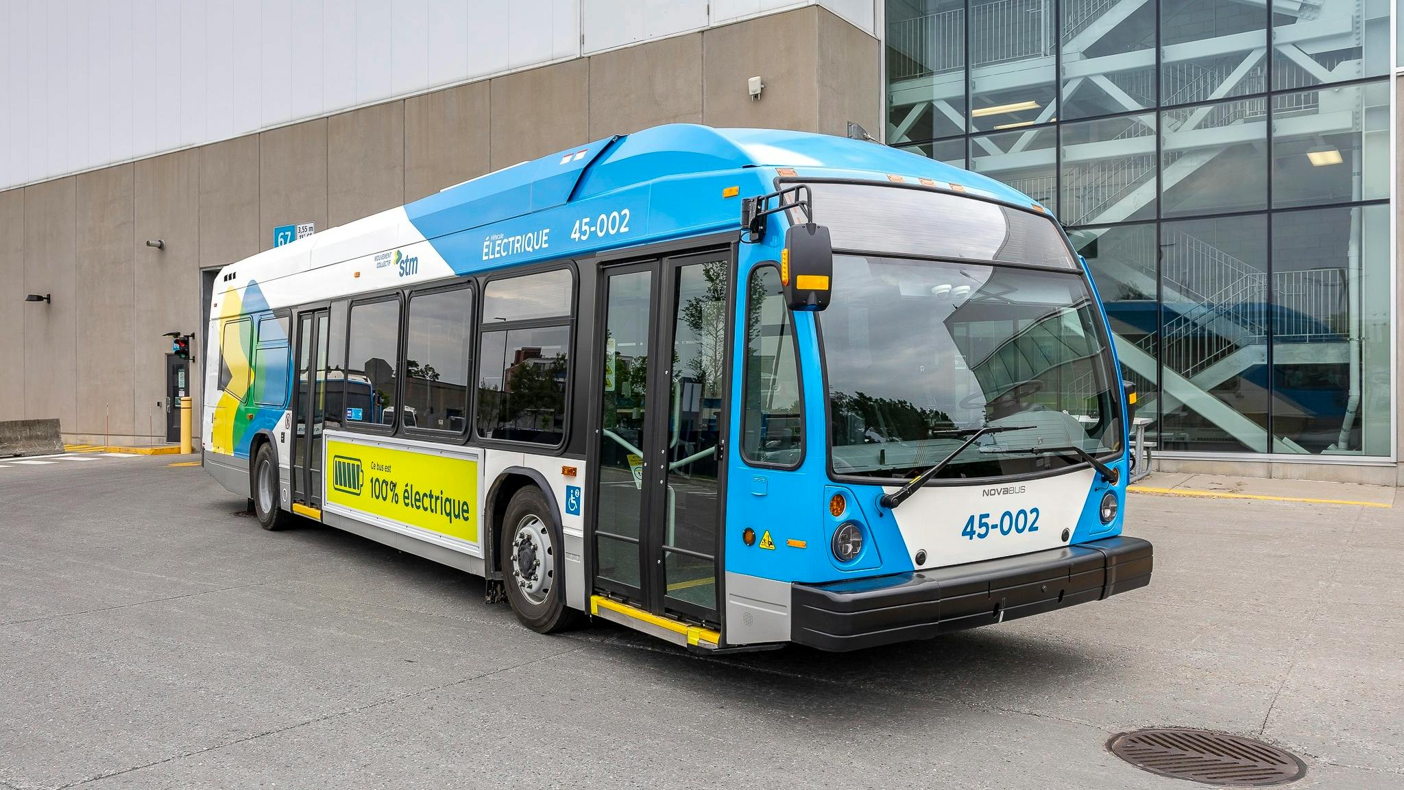 A blue, white and yellow Soci&eacute;t&eacute; de transport de Montr&eacute;al Nova Bus battery-electric bus, numbered 45-002 and marked 'V&eacute;hicule &Eacute;lectrique' and 'Ce bus est 100% &eacute;lectrique,' sits parked outside a transit garage with reflective glass facade.