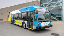 A blue, white and yellow Société de transport de Montréal Nova Bus battery-electric bus, numbered 45-002 and marked 'Véhicule Électrique' and 'Ce bus est 100% électrique,' sits parked outside a transit garage with reflective glass facade. A blue, white and yellow Société de transport de Montréal Nova Bus battery-electric bus, numbered 45-002 and marked 'Véhicule Électrique' and 'Ce bus est 100% électrique,' sits parked outside a transit garage with reflective glass facade.