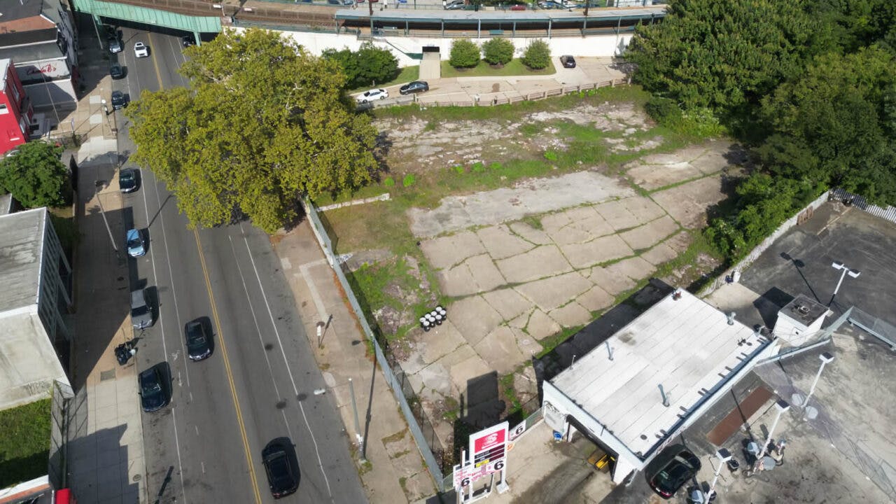 An aerial view of a vacant, cracked-pavement lot adjacent to the Germantown SEPTA Regional Rail station, with a large tree at one corner, neighboring commercial buildings and an elevated rail platform visible in the background.