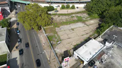 An aerial view of a vacant, cracked-pavement lot adjacent to the Germantown SEPTA Regional Rail station, with a large tree at one corner, neighboring commercial buildings and an elevated rail platform visible in the background.