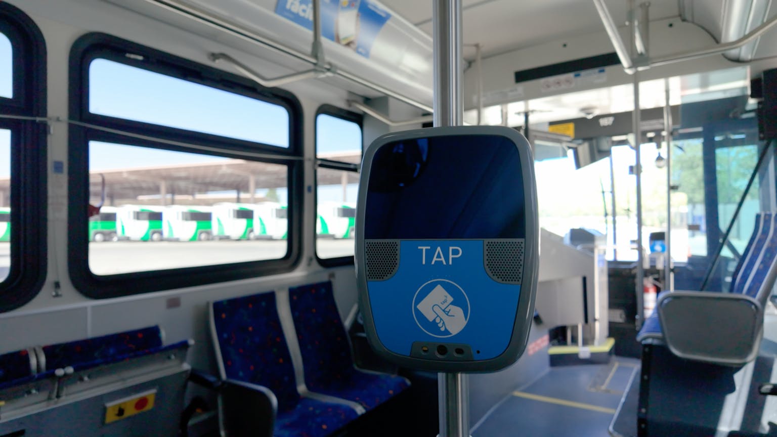 A blue TAP contactless fare reader mounted on a stanchion inside a transit bus interior, with patterned blue seats, a row of windows overlooking a fleet of buses parked outside and the driver's compartment visible at right.