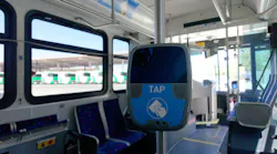 A blue TAP contactless fare reader mounted on a stanchion inside a transit bus interior, with patterned blue seats, a row of windows overlooking a fleet of buses parked outside and the driver's compartment visible at right. A blue TAP contactless fare reader mounted on a stanchion inside a transit bus interior, with patterned blue seats, a row of windows overlooking a fleet of buses parked outside and the driver's compartment visible at right.