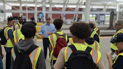 A man in a blue shirt and ball cap speaks to a group of students wearing high-visibility safety vests and backpacks on a Denver Union Station platform, with the station's distinctive white canopy structure overhead and other passengers visible in the background. A man in a blue shirt and ball cap speaks to a group of students wearing high-visibility safety vests and backpacks on a Denver Union Station platform, with the station's distinctive white canopy structure overhead and other passengers visible in the background.