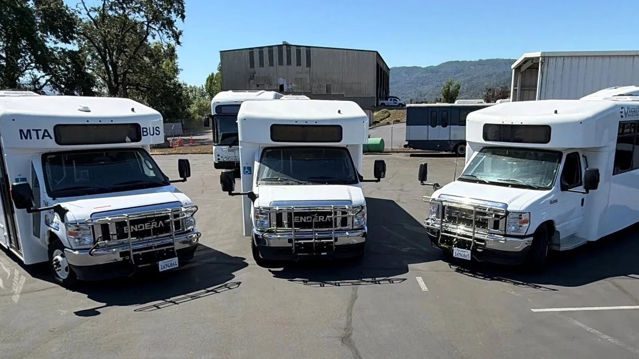Three white Mendocino Transit Authority Endera battery-electric cutaway buses sit parked side by side in a transit yard, with additional buses, agency buildings and tree-covered hills visible in the background.