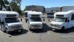 Three white Mendocino Transit Authority Endera battery-electric cutaway buses sit parked side by side in a transit yard, with additional buses, agency buildings and tree-covered hills visible in the background. Three white Mendocino Transit Authority Endera battery-electric cutaway buses sit parked side by side in a transit yard, with additional buses, agency buildings and tree-covered hills visible in the background.