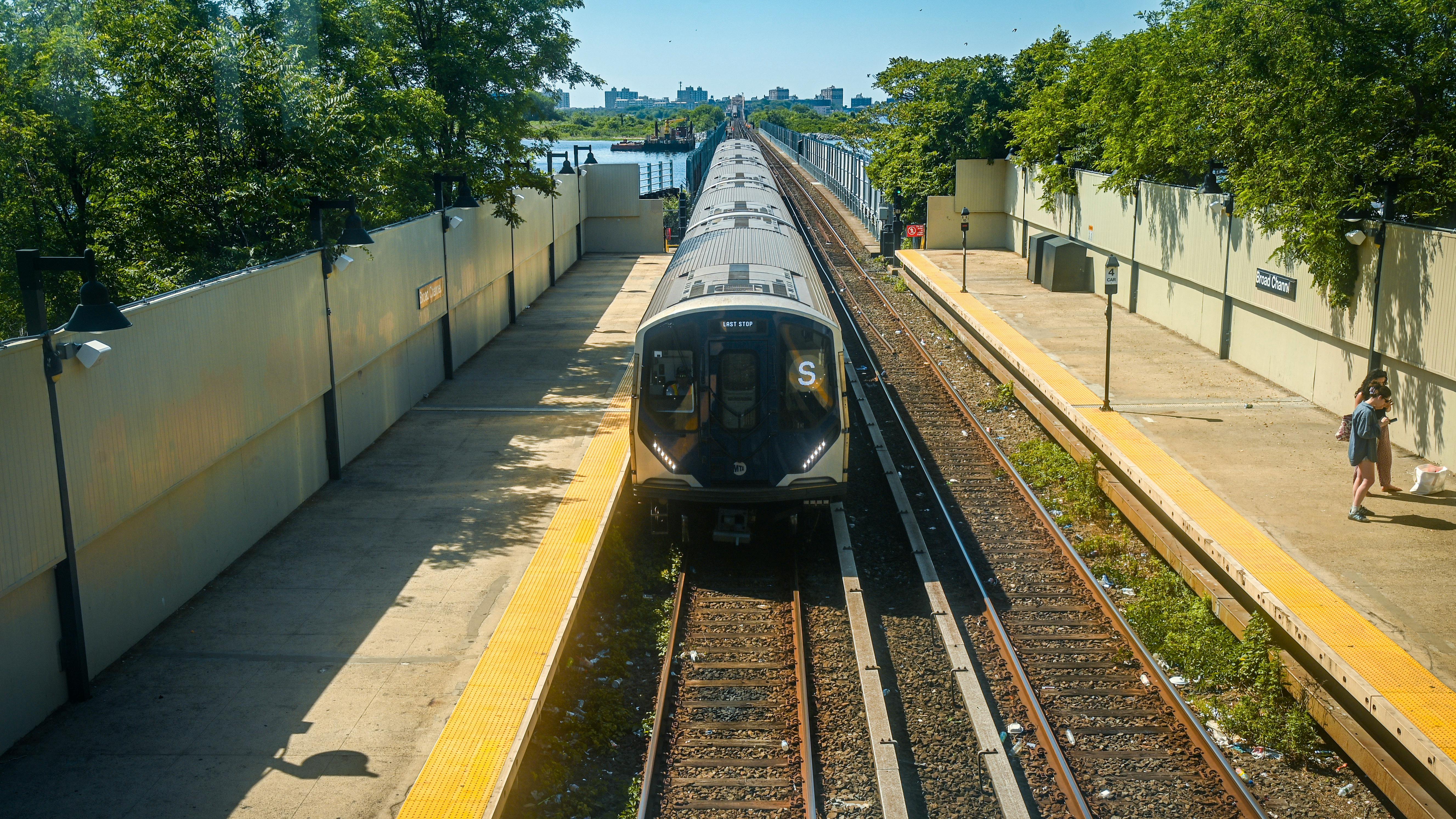 A new MTA R211 subway train enters Broad Channel station on the elevated Rockaway line as part of the Rockaway Park Shuttle's rolling stock upgrade, with Jamaica Bay marshland and a distant skyline behind.