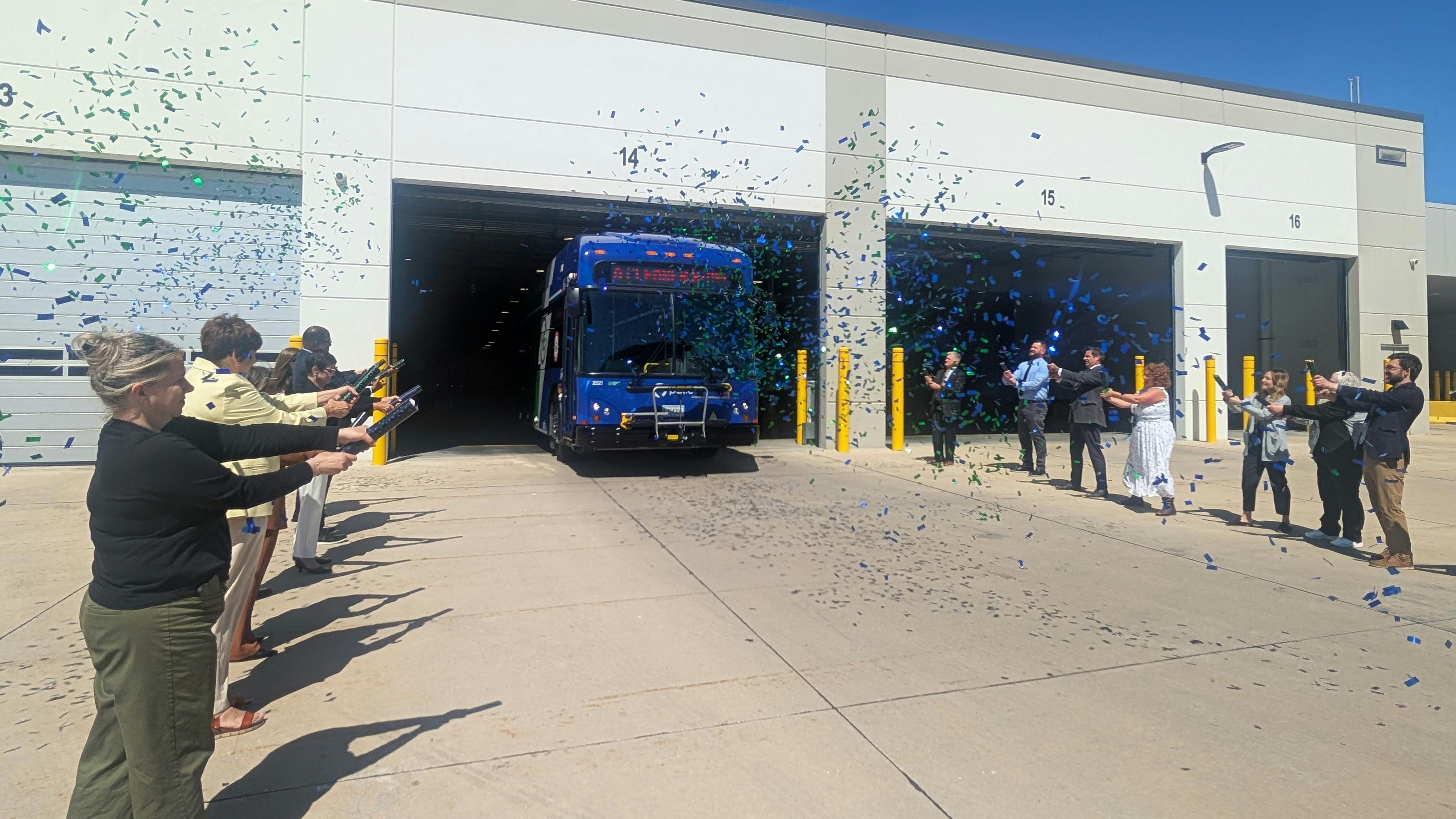 Officials and staff fire blue and green confetti cannons from both sides of a transit garage driveway as a new blue hybrid bus rolls out of a bay marked '14,' with bright sunlight and clear blue sky overhead.