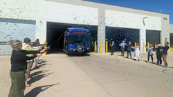 Officials and staff fire blue and green confetti cannons from both sides of a transit garage driveway as a new blue hybrid bus rolls out of a bay marked '14,' with bright sunlight and clear blue sky overhead. Officials and staff fire blue and green confetti cannons from both sides of a transit garage driveway as a new blue hybrid bus rolls out of a bay marked '14,' with bright sunlight and clear blue sky overhead.