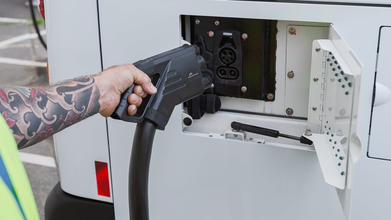A worker in a high-visibility vest plugs a CCS charging connector into the receptacle of a white BC Transit battery-electric bus, with additional buses lined up in the background.