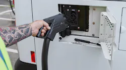 A worker in a high-visibility vest plugs a CCS charging connector into the receptacle of a white BC Transit battery-electric bus, with additional buses lined up in the background.