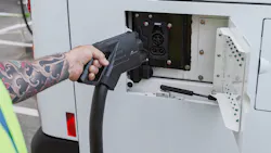 A worker in a high-visibility vest plugs a CCS charging connector into the receptacle of a white BC Transit battery-electric bus, with additional buses lined up in the background. A worker in a high-visibility vest plugs a CCS charging connector into the receptacle of a white BC Transit battery-electric bus, with additional buses lined up in the background.