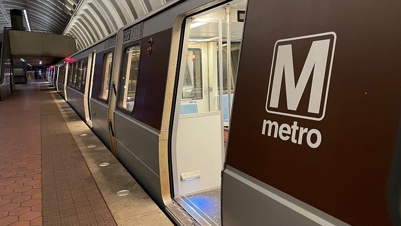 A Washington Metropolitan Area Transit Authority Metrorail train sits at an underground platform with an open door revealing the brightly lit interior, with the agency's 'M metro' wordmark on the brown side panels and the station's distinctive coffered concrete vault ceiling overhead.