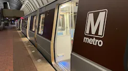 A Washington Metropolitan Area Transit Authority Metrorail train sits at an underground platform with an open door revealing the brightly lit interior, with the agency's 'M metro' wordmark on the brown side panels and the station's distinctive coffered concrete vault ceiling overhead.