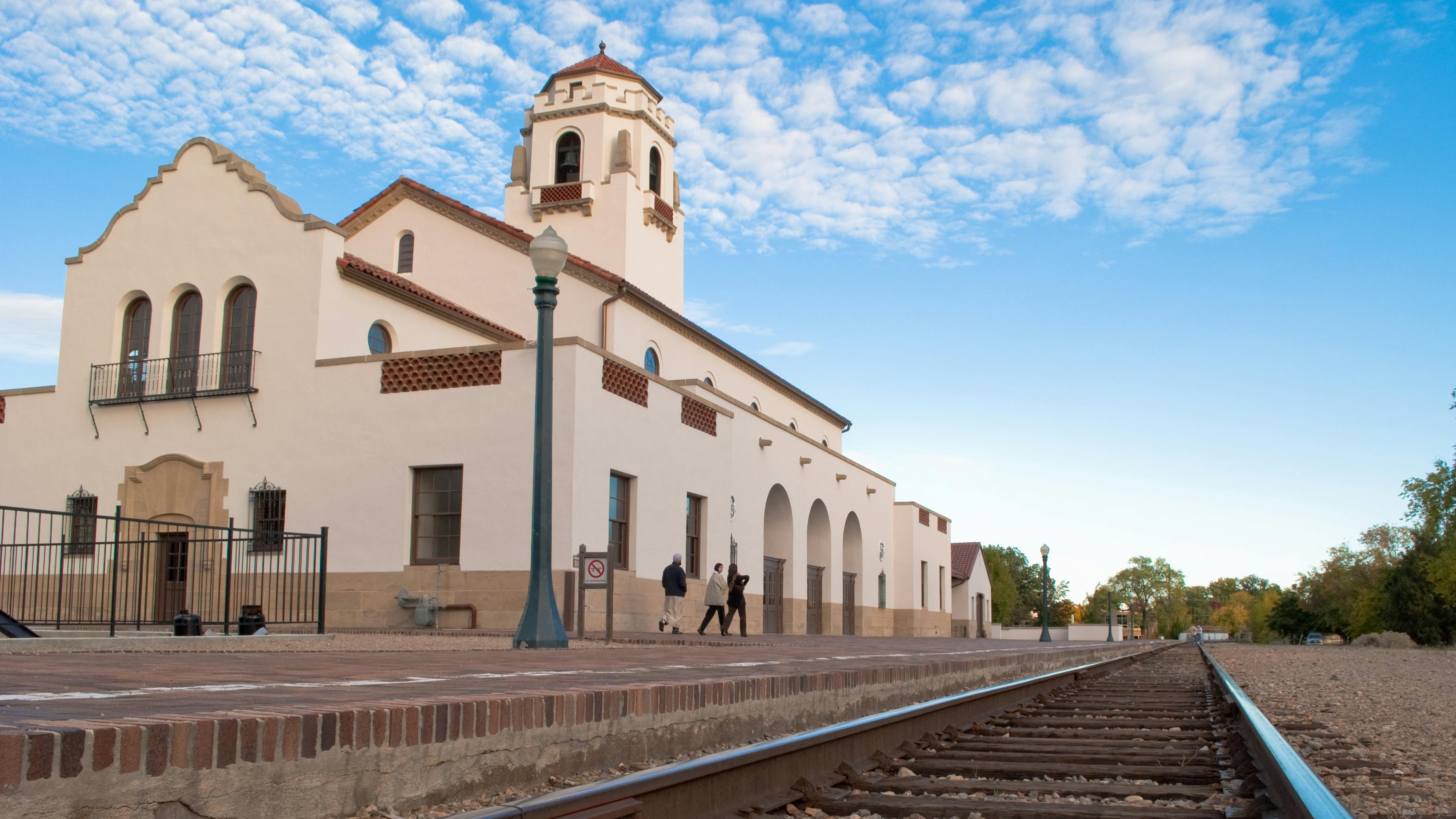 The historic Boise Depot, a Spanish Mission Revival-style train station with a stucco facade, red tile roof and prominent bell tower, viewed from the platform level with rail tracks running alongside and pedestrians walking past the arched entry.