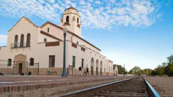 The historic Boise Depot, a Spanish Mission Revival-style train station with a stucco facade, red tile roof and prominent bell tower, viewed from the platform level with rail tracks running alongside and pedestrians walking past the arched entry. The historic Boise Depot, a Spanish Mission Revival-style train station with a stucco facade, red tile roof and prominent bell tower, viewed from the platform level with rail tracks running alongside and pedestrians walking past the arched entry.