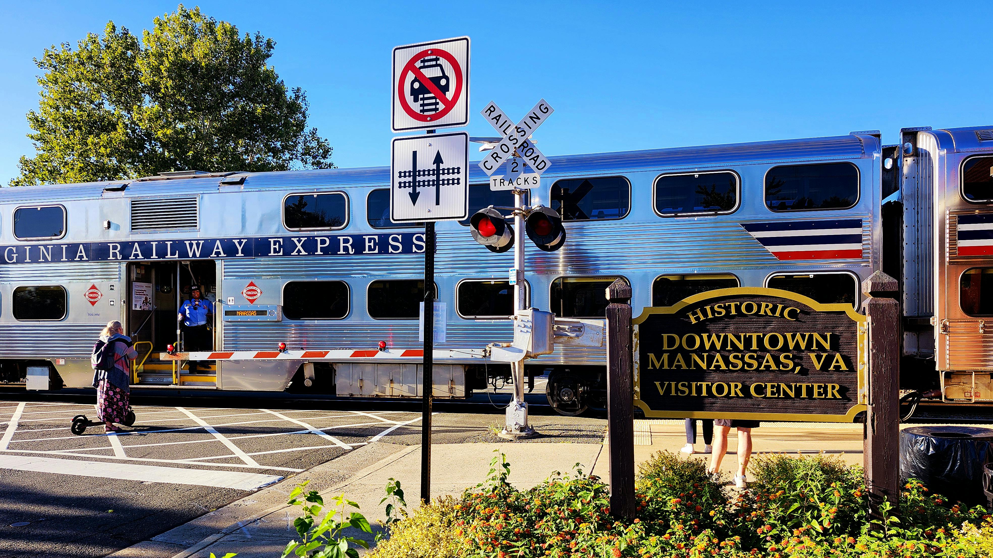 A silver Virginia Railway Express bilevel commuter rail car with red, white and blue striping sits at a grade crossing with a conductor visible in the open doorway, with a 'Historic Downtown Manassas, VA Visitor Center' sign and railroad crossing signals in the foreground.